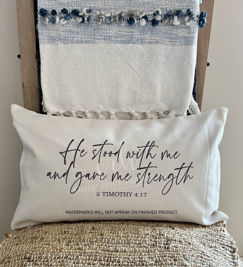 Może przedstawiać: Biała poduszka z czarnym tekstem, kt&oacute;ry brzmi "He stood with me and gave me strength 2 Timothy 4:17". Poduszka leży na plecionym krześle.