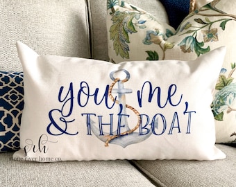 以下が含まれることがあります： 白いクッションに、青い文字で「you & me, & the boat」と書いてあり、青い水彩の錨と茶色のロープが描かれています。クッションは薄い灰色のソファの上に置かれています。