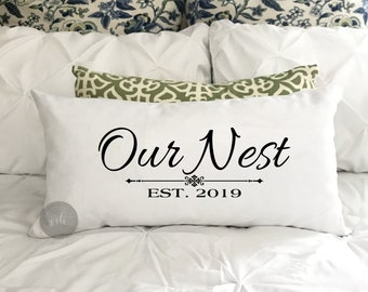 May include: White throw pillow with black text that reads "Our Nest Est. 2019". The pillow is on a white bed with other pillows.