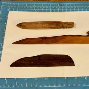 Puede incluir: Tres abridores de cartas de madera en diferentes tonos de marrón. El superior es marrón claro con detalles tallados. El del medio es marrón medio con un nudo. El inferior es marrón oscuro, de forma curva.