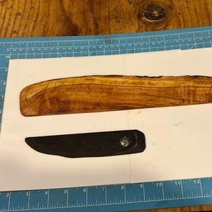 May include: A handcrafted wooden knife handle with a dark, rich wood grain. A black, curved knife blade is positioned below the handle. The items are on a white surface with a blue grid background, suggesting a craft or woodworking project.