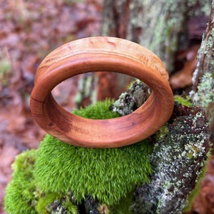 May include: A handcrafted wooden bracelet with a natural, warm brown color. The bracelet is circular and appears to be made from a single piece of wood. The wood grain is visible, adding to its rustic charm. The bracelet is displayed on a bed of green moss.