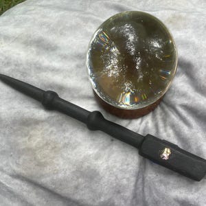 Peut inclure: Une baguette en bois noir avec une pointe et une poignée décorative, à côté d'une boule de cristal transparente sur une base en bois. La boule de cristal présente des motifs internes et repose sur une surface gris clair.