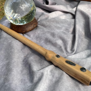 May include: A handcrafted wooden wand with a light brown finish, featuring dark accents and a tapered design. The wand has a smooth, polished appearance and is approximately 14 inches long. It is a magical item, perfect for cosplay or display.