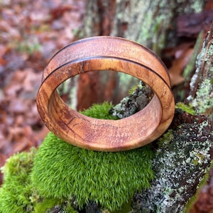 May include: A handcrafted wooden ring with a natural wood grain pattern. The ring is light brown with a dark brown accent line. The ring is set against a backdrop of green moss and a tree branch.