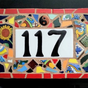 May include: A rectangular mosaic house number sign with the number 117 in black on a white background. The sign is framed with red brick and colorful mosaic tiles, including a sunflower and a yin-yang symbol.