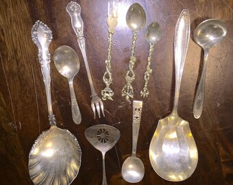 May include: A collection of ten silver spoons and serving spoons, including a slotted spoon, a ladle, and a spoon with a decorative shell-shaped bowl. The spoons are arranged on a wooden surface.