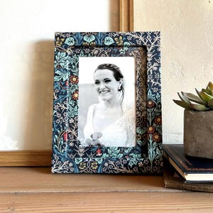May include: A black picture frame with a floral pattern in shades of blue, green, yellow, and red. The frame is holding a black and white photo of a woman smiling.