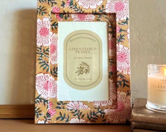 May include: A floral patterned picture frame with a gold background and pink flowers. The frame has a white mat and a gold label that reads "StoreHouse405 Upholstered Frames Artisan Made in Oklahoma".