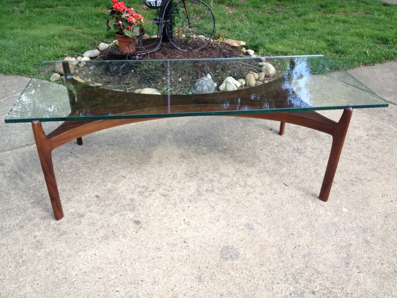 K&ouml;nnte beinhalten: Ein moderner Glas- und Holz-Couchtisch im Mid-Century-Stil mit einer geschwungenen Glasplatte und vier Holzbeinen. Der Tisch ist aus dunklem Holz gefertigt und hat eine nat&uuml;rliche Oberfl&auml;che.