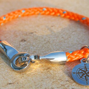 May include: Orange and blue rope bracelet with a silver compass charm. The compass charm has the letters N, S, E, and W engraved on it.