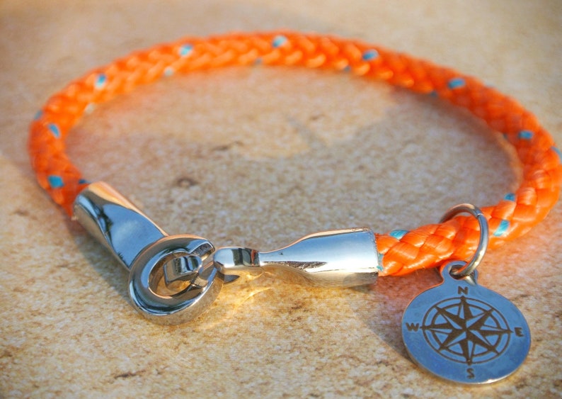 May include: Orange and blue rope bracelet with a silver compass charm. The compass charm has the letters N, S, E, and W engraved on it.