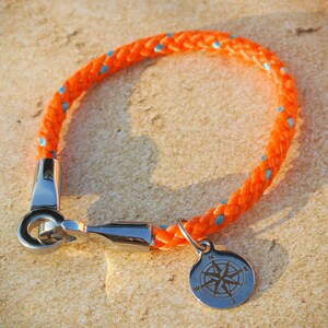 May include: Orange and blue rope bracelet with a silver compass charm. The compass charm has a silver ring and a compass rose design.