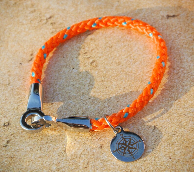 May include: Orange and blue rope bracelet with a silver compass charm. The compass charm has a silver ring and a compass rose design.