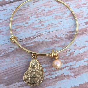 Puede incluir: Pulsera de brazalete dorada con un dije de concha de ostra y una pequeña perla redonda de color rosa claro. El dije de concha de ostra tiene crestas detalladas. La pulsera se muestra sobre una superficie de madera desgastada azul claro y blanca.