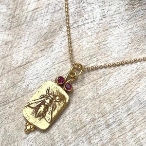 Puede incluir: Collar de oro con un colgante rectangular que presenta un diseño de abeja y dos pequeñas piedras preciosas rosas.