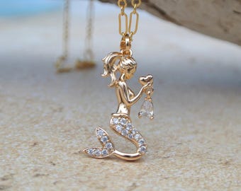 May include: Gold-colored mermaid pendant necklace. The mermaid holds a heart and a teardrop-shaped gem. The mermaid's tail is encrusted with small, clear gemstones. The necklace chain is also gold-colored.