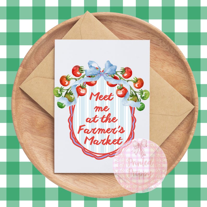 Op de afbeelding: Een wenskaart met de tekst "Meet me at the Farmer's Market" omringd door een aquarel illustratie van tomaten en een blauwe strik. De kaart ligt op een houten dienblad met een bruine envelop, tegen een groen-witte geruite achtergrond.