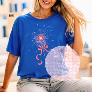 K&ouml;nnte beinhalten: Ein blaues T-Shirt mit einem roten, wei&szlig;en und blauen Feuerwerksk&ouml;rper-Motiv mit einer roten Schleife. Der Text "Printed Preppy" ist im Hintergrund sichtbar.