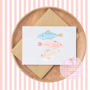 Puede incluir: Una tarjeta con una ilustraci&oacute;n de acuarela de tres peces. Los peces son azules, rosas y blancos. La tarjeta est&aacute; en un plato de madera con un sobre marr&oacute;n.