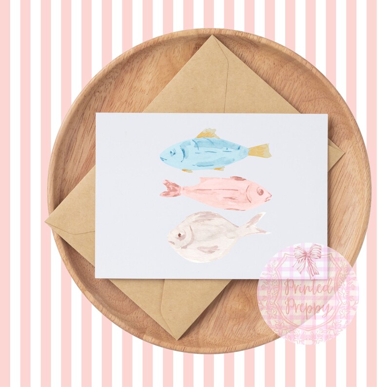 Puede incluir: Una tarjeta con una ilustraci&oacute;n de acuarela de tres peces. Los peces son azules, rosas y blancos. La tarjeta est&aacute; en un plato de madera con un sobre marr&oacute;n.