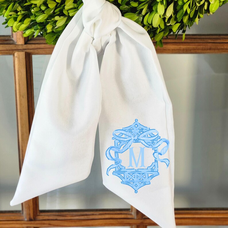 May include: White scarf with a light blue embroidered design featuring the letter "M" inside a decorative frame. The scarf is tied in a knot at the top and hangs down. The design includes ribbons and geometric patterns.