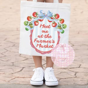 Op de afbeelding: Witte draagtas met een aquarelontwerp van rode tomaten, groene takjes en een blauwe strik. De tekst "Meet me at the Farmer's Market" wordt weergegeven in een decoratief kader. De tas wordt gedragen door een persoon die witte sneakers draagt.