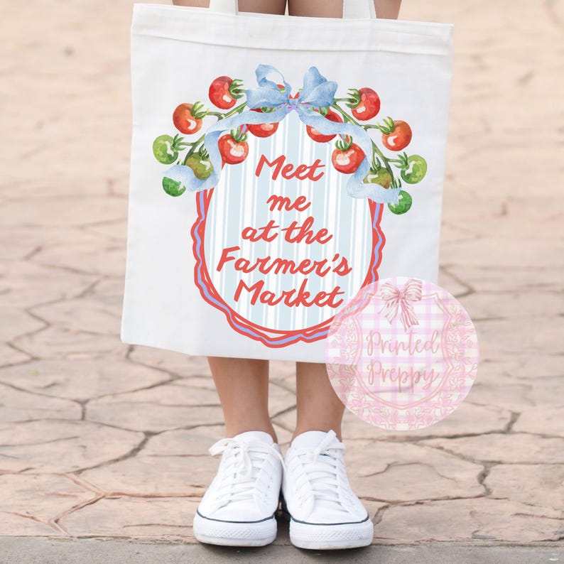 Op de afbeelding: Witte draagtas met een aquarelontwerp van rode tomaten, groene takjes en een blauwe strik. De tekst "Meet me at the Farmer's Market" wordt weergegeven in een decoratief kader. De tas wordt gedragen door een persoon die witte sneakers draagt.