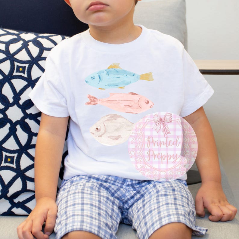 Puede incluir: Una camiseta blanca con tres dise&ntilde;os de peces de acuarela. Los peces son azules, rosas y beige. La camiseta tiene el texto "Printed Preppy" sobre un fondo de cuadros rosa y blanco.