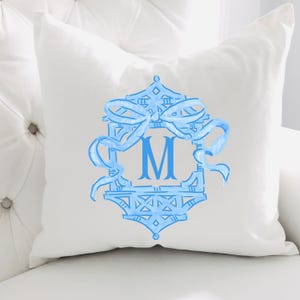 May include: White square throw pillow with a light blue monogram design. The design features a decorative frame with a bow and the letter "M" in the center. The pillow is on a white surface.