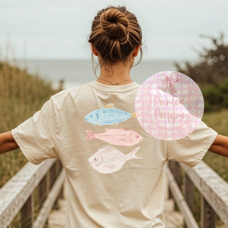 Puede incluir: Una camiseta beige claro con un gr&aacute;fico de tres peces en tonos de azul, rosa y blanco. Los peces est&aacute;n dispuestos verticalmente, con el pez m&aacute;s grande en la parte superior y el pez m&aacute;s peque&ntilde;o en la parte inferior. La camiseta tiene cuello redondo y mangas cortas. El texto "Printed Preppy" es visible sobre un fondo de cuadros rosa y blanco.