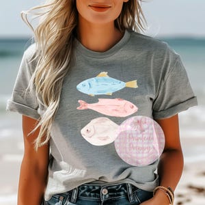 Puede incluir: Una camiseta gris con un dise&ntilde;o de tres peces de acuarela. Los peces son azules, rosas y blancos. La camiseta tiene cuello redondo y mangas cortas.