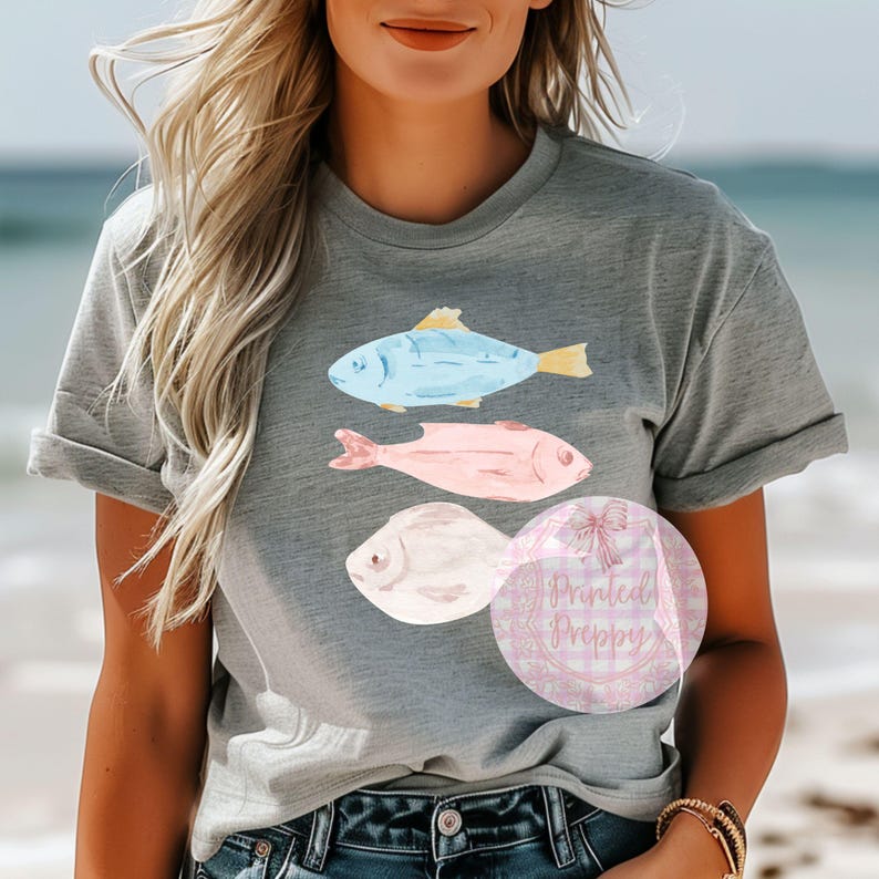 Puede incluir: Una camiseta gris con un dise&ntilde;o de tres peces de acuarela. Los peces son azules, rosas y blancos. La camiseta tiene cuello redondo y mangas cortas.