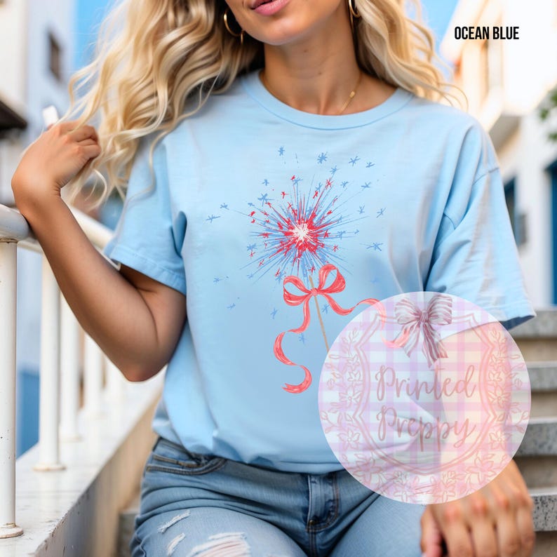 K&ouml;nnte beinhalten: Ein hellblaues T-Shirt mit einem rot-wei&szlig;-blauen Feuerwerksdesign mit einer rosa Schleife. Der Text "Printed Preppy" ist in der rechten unteren Ecke des Bildes sichtbar.