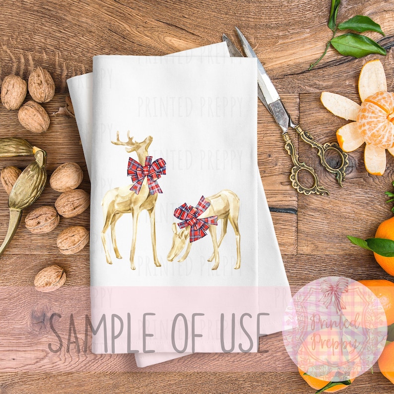 Puede incluir: Toalla decorativa blanca con dos ciervos dorados con lazos de cuadros rojos y negros. La toalla tiene el texto "SAMPLE OF USE" impreso. La toalla est&aacute; sobre una superficie de madera con nueces y tijeras.