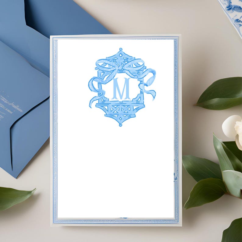 May include: White stationery with a light blue border and a light blue monogram. The monogram features the letter "M" within a decorative frame with ribbons. A matching blue envelope is visible in the background.