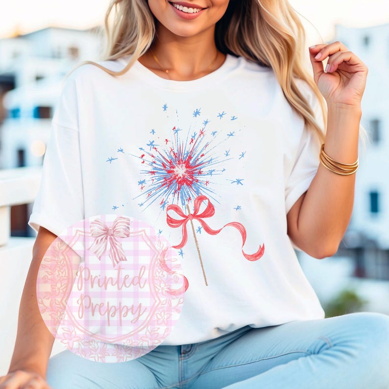 K&ouml;nnte beinhalten: Ein wei&szlig;es T-Shirt mit einem rot-wei&szlig;-blauen Feuerwerksdesign. Das Feuerwerk ist mit einer roten Schleife verziert. Das Shirt tr&auml;gt die Worte "Printed Preppy" in Pink auf einem rosa-wei&szlig; karierten Hintergrund.