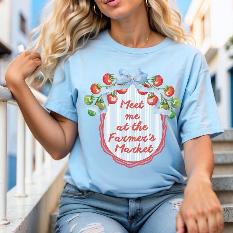 Op de afbeelding: Lichtblauw T-shirt met de tekst "Meet me at the Farmer's Market". De afbeelding toont een krans van rode en groene tomaten en een blauwe strik. Het T-shirt is gemaakt van zachte stof.
