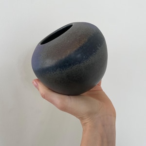 May include: A dark gray ceramic vase with a unique, rounded shape and an angled opening. The vase has a textured surface with a dark blue band across the middle. The vase is held in a hand against a white background.