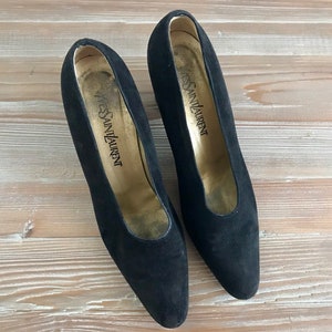 Peut inclure: Une paire de escarpins noirs en daim &agrave; talons hauts et &agrave; bout pointu. Les chaussures ont une semelle int&eacute;rieure dor&eacute;e avec le texte "Yves Saint Laurent" imprim&eacute; dessus.