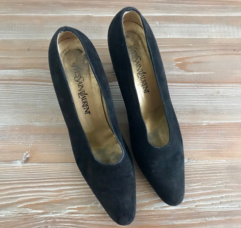 Peut inclure: Une paire de escarpins noirs en daim &agrave; talons hauts et &agrave; bout pointu. Les chaussures ont une semelle int&eacute;rieure dor&eacute;e avec le texte "Yves Saint Laurent" imprim&eacute; dessus.