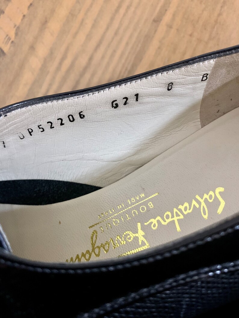 Pu&ograve; includere: Primo piano dell'interno di una scarpa in pelle nera con il testo "Salvatore Ferragamo Boutique Made in Italy" e i numeri "UP 52206 621 8 B" stampati sulla soletta.