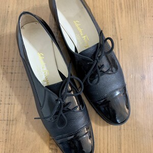 Pu&ograve; includere: Coppia di scarpe stringate in pelle nera con punta in vernice. Le scarpe hanno un tacco basso e sono realizzate da Salvatore Ferragamo.