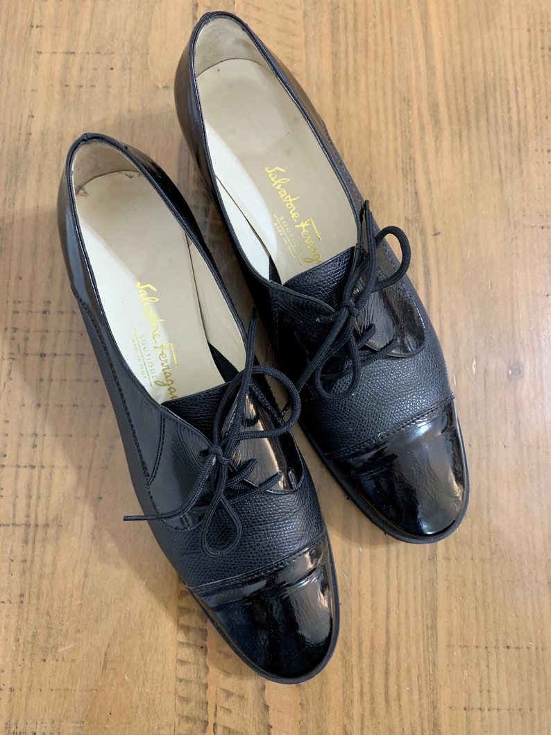 Pu&ograve; includere: Coppia di scarpe stringate in pelle nera con punta in vernice. Le scarpe hanno un tacco basso e sono realizzate da Salvatore Ferragamo.