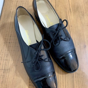 Pu&ograve; includere: Un paio di scarpe stringate in pelle nera con punta in vernice. Le scarpe hanno un design classico e sono realizzate da Salvatore Ferragamo.
