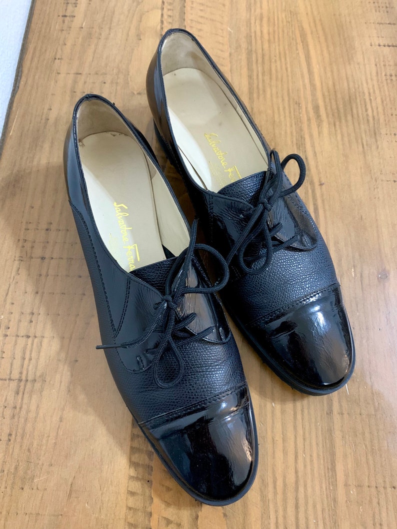 Pu&ograve; includere: Un paio di scarpe stringate in pelle nera con punta in vernice. Le scarpe hanno un design classico e sono realizzate da Salvatore Ferragamo.
