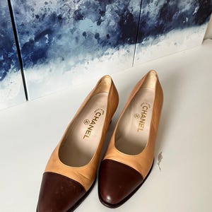 Puede incluir: Un par de zapatos de tacón de cuero color canela y marrón oscuro con puntera redondeada. Las plantillas interiores presentan el logotipo de Chanel y el texto Made in Italy. Estos zapatos tienen un tacón bajo y un acabado de cuero liso.