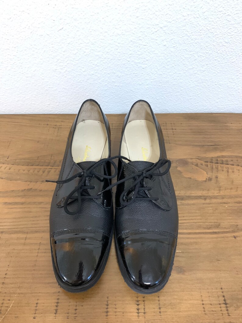 Pu&ograve; includere: Un paio di scarpe stringate in pelle nera con punta in vernice.