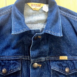 May include: A blue denim jacket with a brown leather label that reads "Rustler" and "S" in white lettering. The jacket has two front pockets and a button closure.