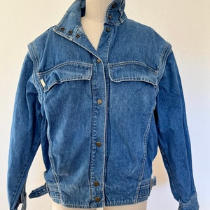 Könnte beinhalten: Eine blaue Jeansjacke mit Stehkragen, Knopfleiste und zwei Pattentaschen. Die Jacke hat einen Gürtelbund und lange Ärmel.